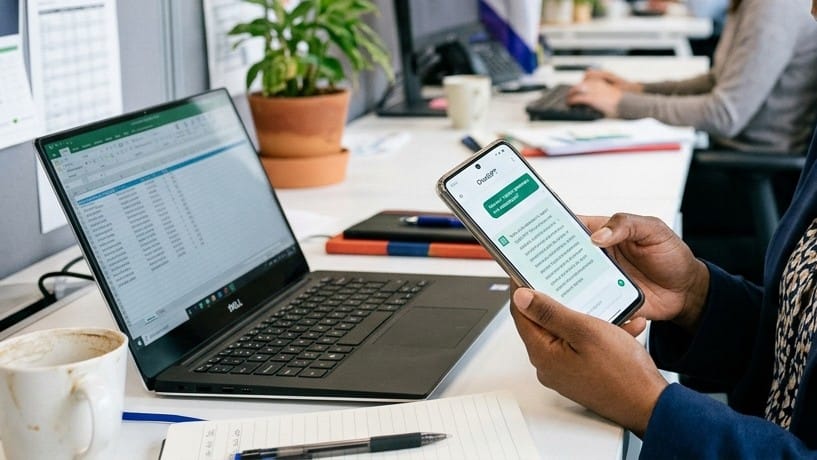 Mão segurando celular com conversa de IA aberta, notebook com planilha ao fundo